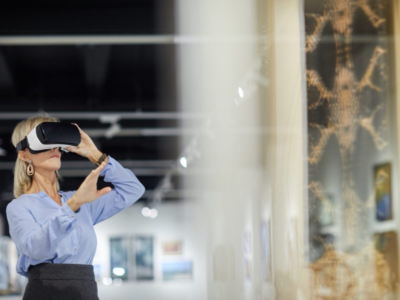 Wide angle portrait of elegant mature woman wearing VR gear while enjoying immersive experience during exhibition in art gallery, copy space